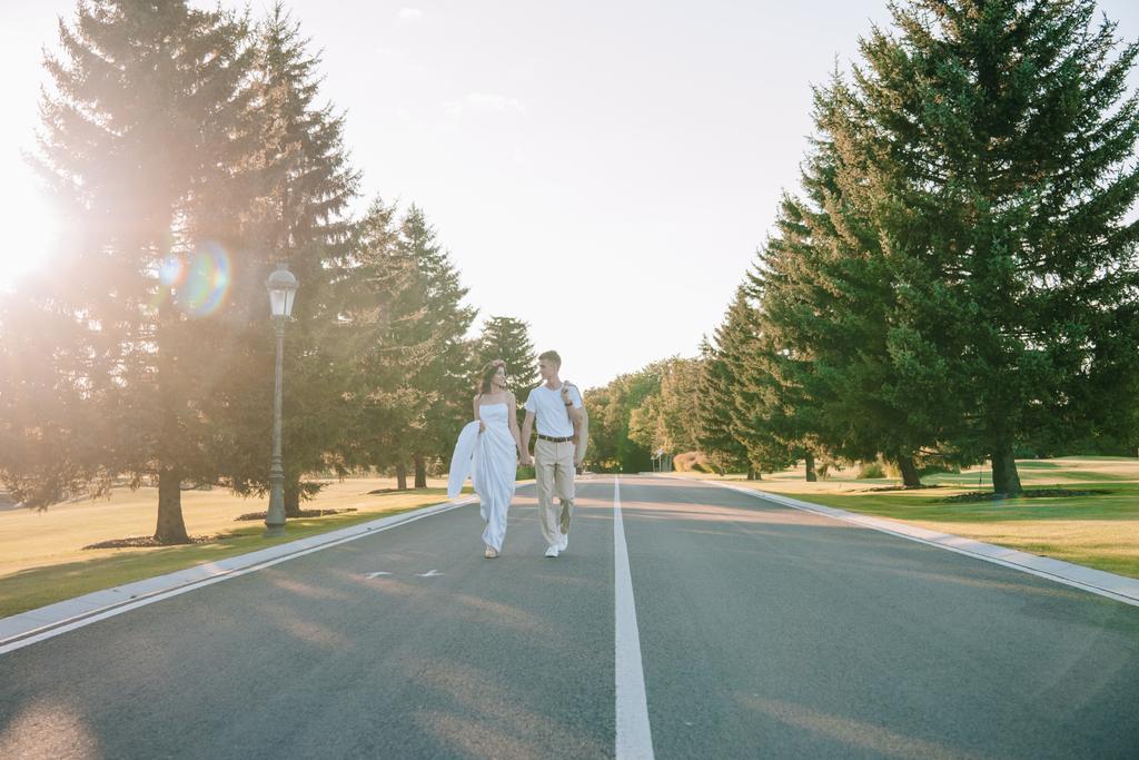 手を繋いでいると、夕暮れ時の公園で一緒に歩きながら笑みを浮かべてお互い若い結婚式のカップル  - 写真・画像