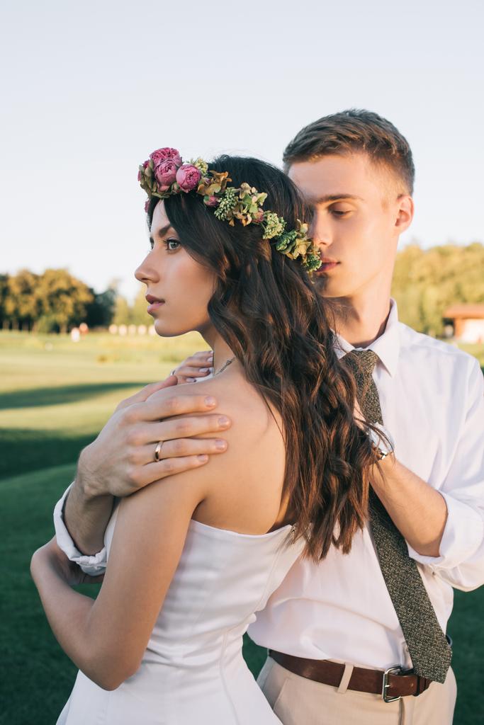 junger Bräutigam umarmt schöne junge Braut in Blumenkranz im Park - Foto, Bild