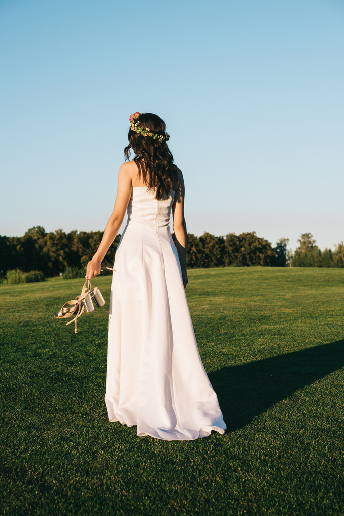 zadní pohled na nabídku mladá nevěsta drží boty a chodit na zeleném trávníku  - Fotografie, Obrázek
