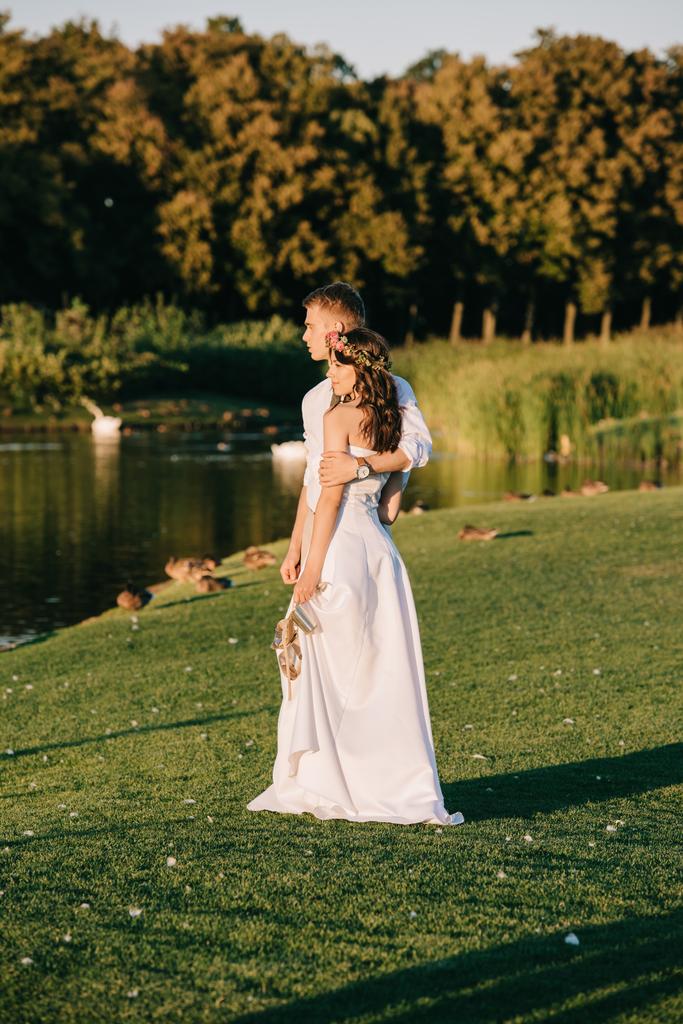 公園の湖の近くに立っている間を受け入れる美しい幸せな若い結婚式のカップル - 写真・画像