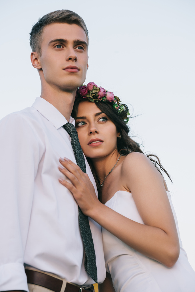 low angle view of beautiful romantic young wedding couple embracing and looking away - Photo, Image