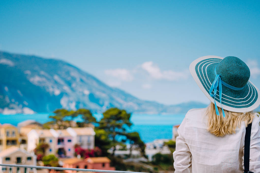 Tourist woman wear blue sunhat and white clothes admire view of colorful tranquil village Assos on sunny day. Stylish female visiting Kefalonia enjoying summer time on Greece travel vacation - Photo, Image