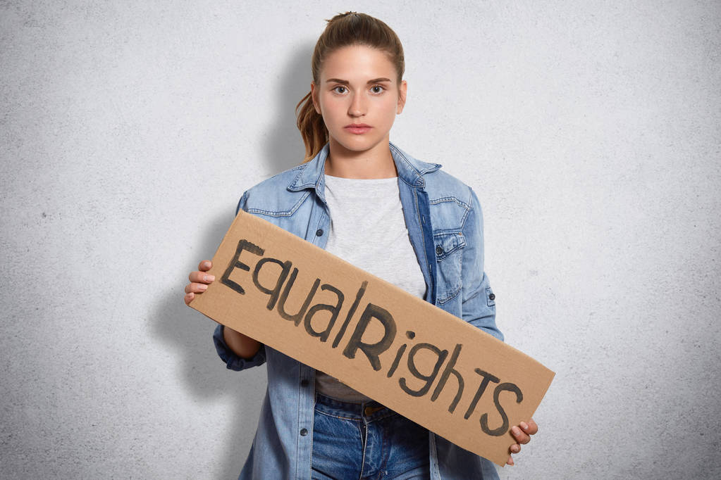 Photo of feminist in denim jacket, holds sign written Equal rights, shows that women have rights and power, isoated over white concrete wall, looks seriously at camera. Womens defense concept - Photo, Image
