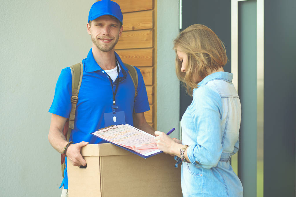 Lachende bezorger in blauw uniform levert pakketdoos aan ontvanger - koerier service concept. Glimlachende bezorger in blauw uniform - Foto, afbeelding
