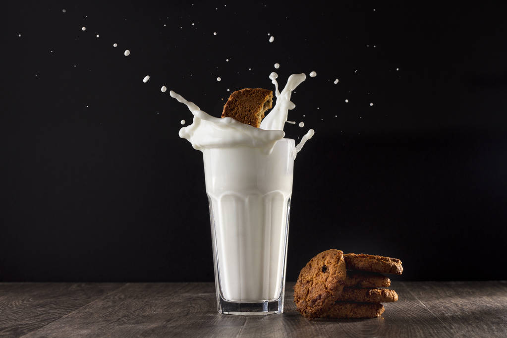 Biscoitos de aveia e copo de leite em uma mesa de madeira em um fundo preto. Um pedaço de biscoito cai num copo
. - Foto, Imagem