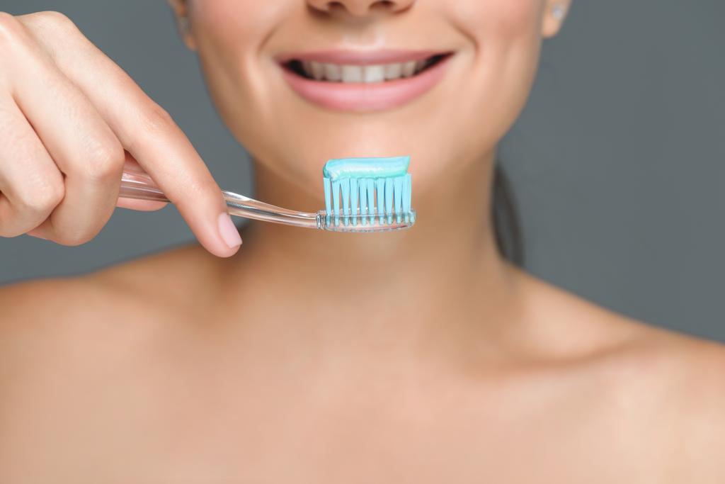 partial view of smiling woman holding tooth brush with tooth paste isolated on grey - Photo, Image