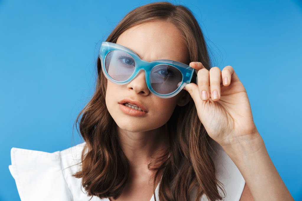 Portrait of a confused young girl in summer clothes wearing sunglasses isolated over blue background - Photo, Image