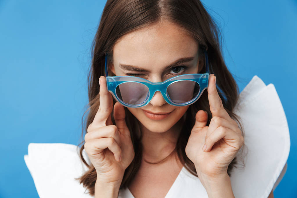 Πορτρέτο του ένα παιχνιδιάρικο νεαρό κορίτσι σε ρούχα καλοκαίρι που αναζητούν πάνω από τα γυαλιά ηλίου που απομονώνονται σε μπλε φόντο - Φωτογραφία, εικόνα