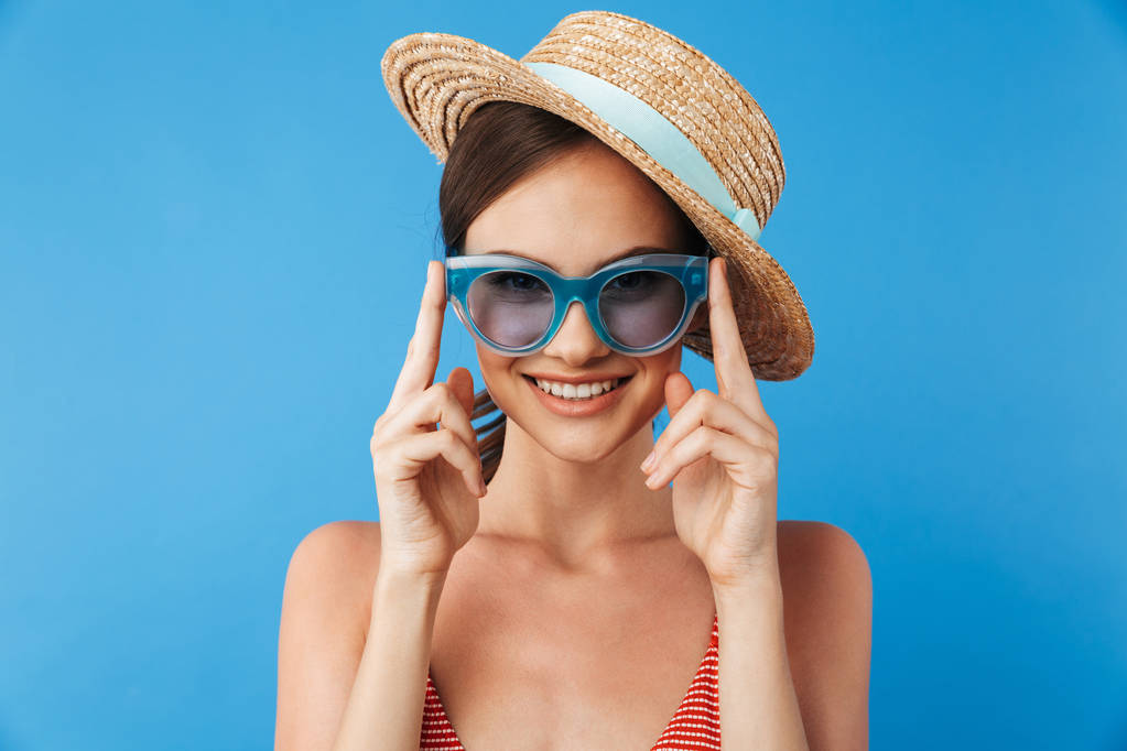 Portréja egy vonzó, fiatal lány a fürdőruhát visel, napszemüveget és nyári kalap pózol, látszó-on fényképezőgép, elszigetelt, felett kék háttér - Fotó, kép