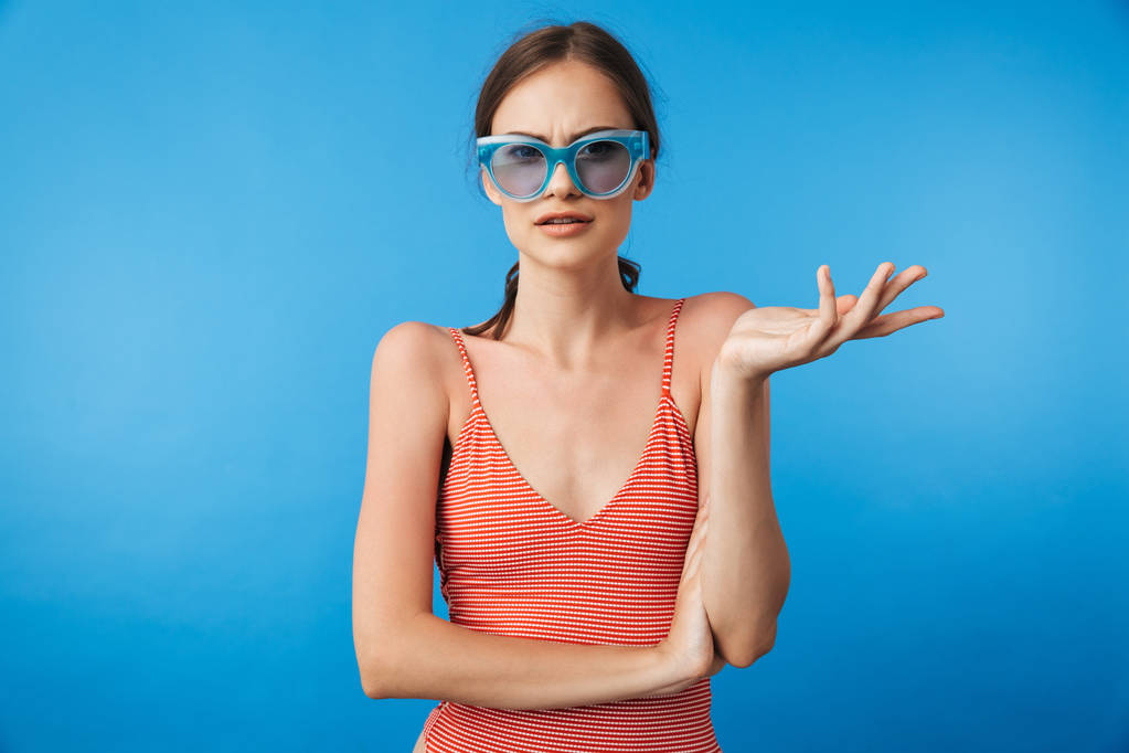 Retrato de una joven confusa en traje de baño con gafas de sol mirando a la cámara aislada sobre fondo azul
 - Foto, Imagen