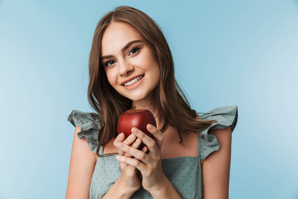 İçinde mavi arka plan üzerinde izole kırmızı elma tutan bir gülümseyen genç kadın portresi - Fotoğraf, Görsel