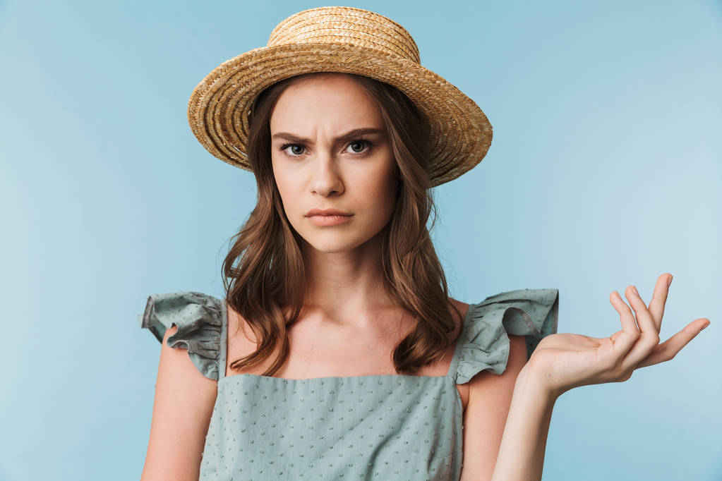 Close up retrato de uma mulher com raiva no vestido e chapéu de verão olhando para a câmera isolada sobre fundo azul
 - Foto, Imagem