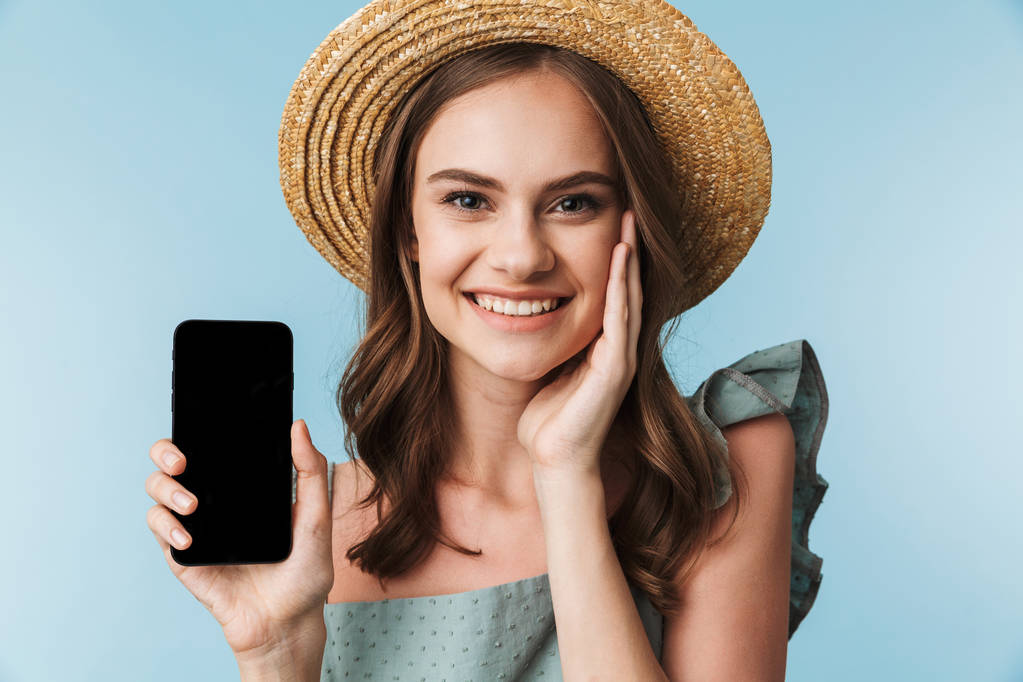 Gros plan portrait d'une jeune femme gaie en robe et chapeau d'été montrant écran vierge téléphone portable isolé sur fond bleu
 - Photo, image