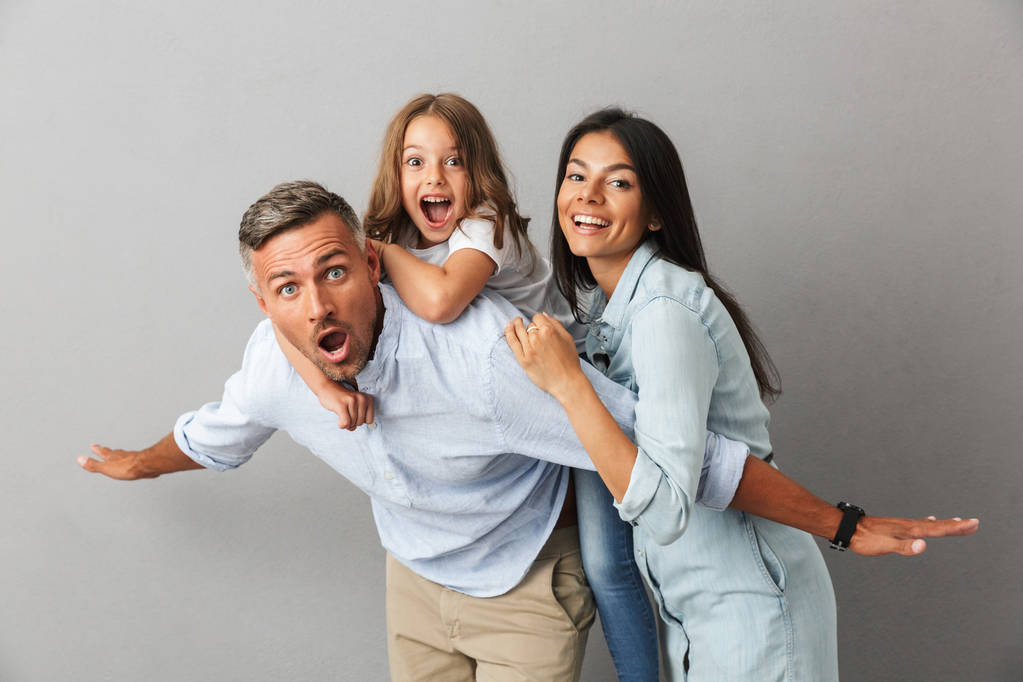 Πορτρέτο του μια χαρούμενη οικογένεια ο πατέρας, η μητέρα, η μικρή κόρη του διασκεδάζοντας μαζί, κρατώντας το κοριτσάκι στην πλάτη του πατέρα απομονωμένη πάνω από το γκρίζο φόντο - Φωτογραφία, εικόνα