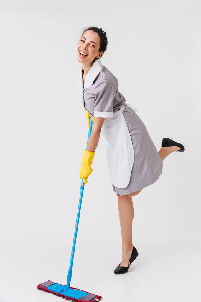 Retrato de larga duración de una joven criada alegre vestida de uniforme usando una fregona aislada sobre fondo blanco
 - Foto, Imagen