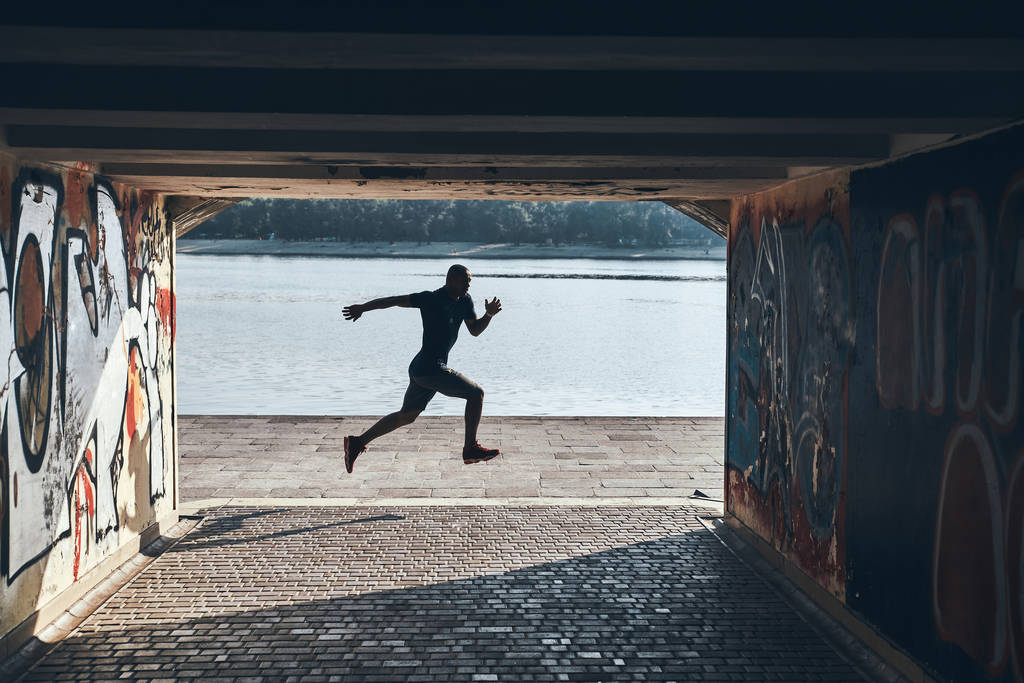 Прыжок в длину Полная длина молодой африканский мужчина в спортивной одежде бег трусцой во время тренировки у реки на открытом воздухе
 - Фото, изображение