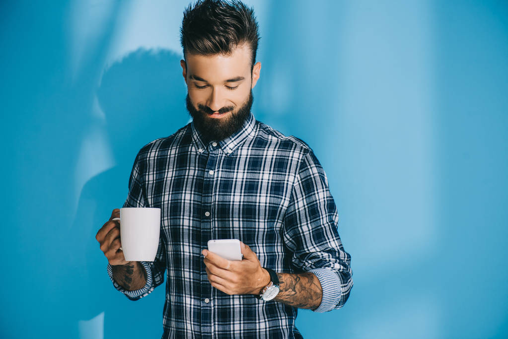 homem barbudo em camisa quadriculada segurando smartphone e xícara de café, em azul
 - Foto, Imagem