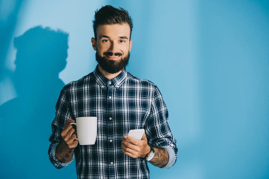 スマート フォンと一杯のコーヒーを保持している格子縞のシャツの男の笑みを浮かべてください。 - 写真・画像