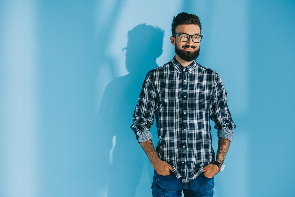 handsome bearded man in checkered shirt standing with hands in pockets, on blue  - Photo, Image