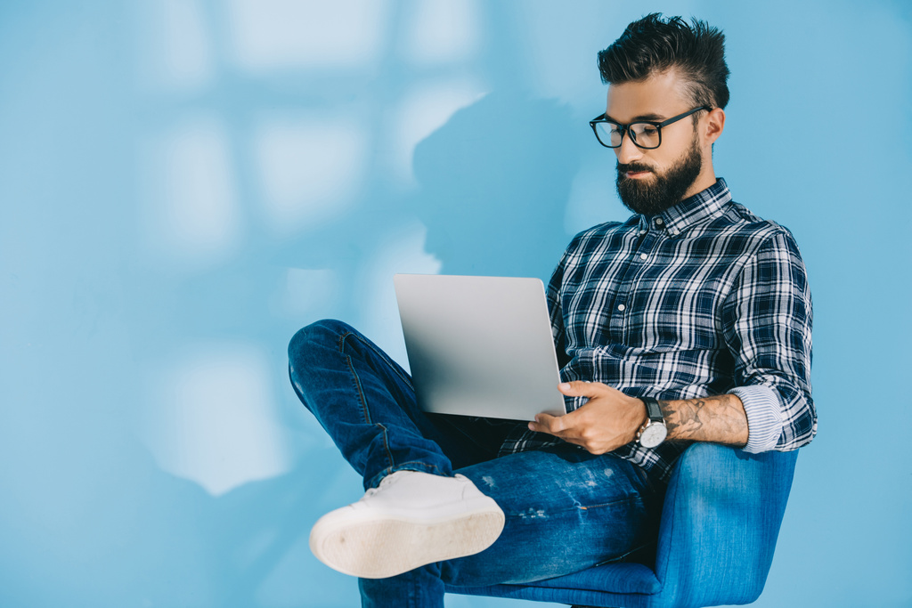 bearded man in checkered shirt using laptop and sitting in armchair, on blue - Photo, Image