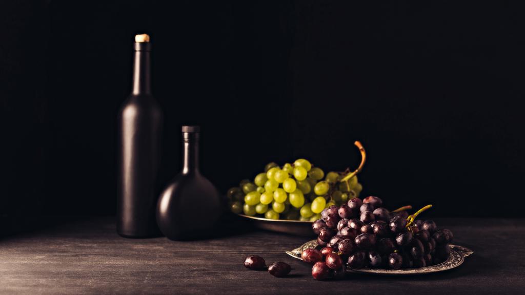köstliche reife rote und weiße Trauben auf Vintage-Tellern und Weinflaschen auf Holztisch auf Schwarz - Foto, Bild