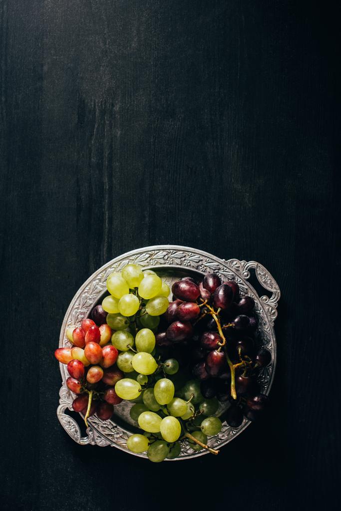 bovenaanzicht van verschillende soorten druiven op vintage plaat op zwarte houten oppervlak - Foto, afbeelding