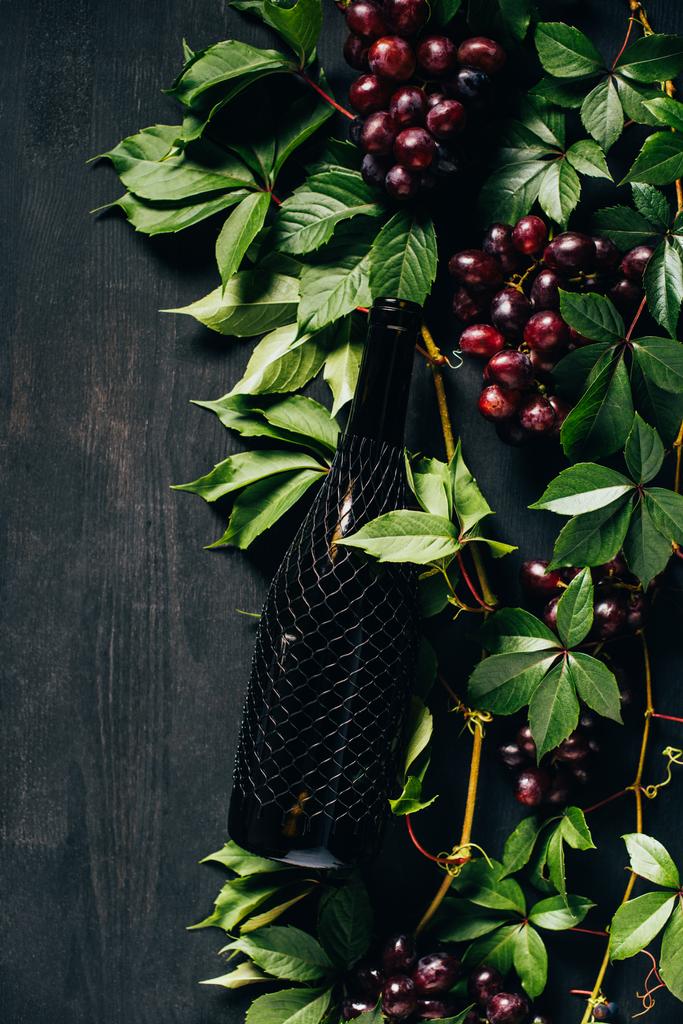黒の木製の背景に新鮮な熟したブドウ、緑の葉とワインのボトルのトップ ビュー   - 写真・画像