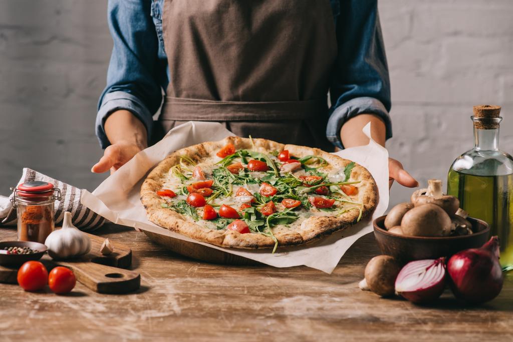 vista parcial de la mujer en delantal sosteniendo pizza cocida en la mesa de madera y los ingredientes
 - Foto, Imagen
