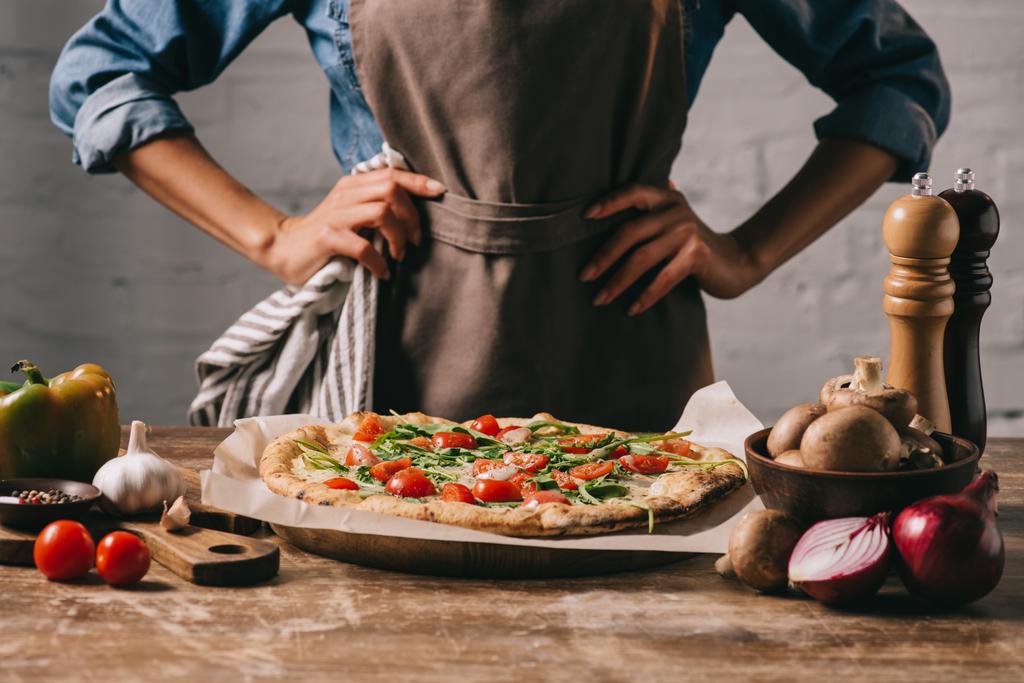 частичный вид женщины в фартуке, стоящей на поверхности с приготовленной пиццей и ингредиентами
 - Фото, изображение