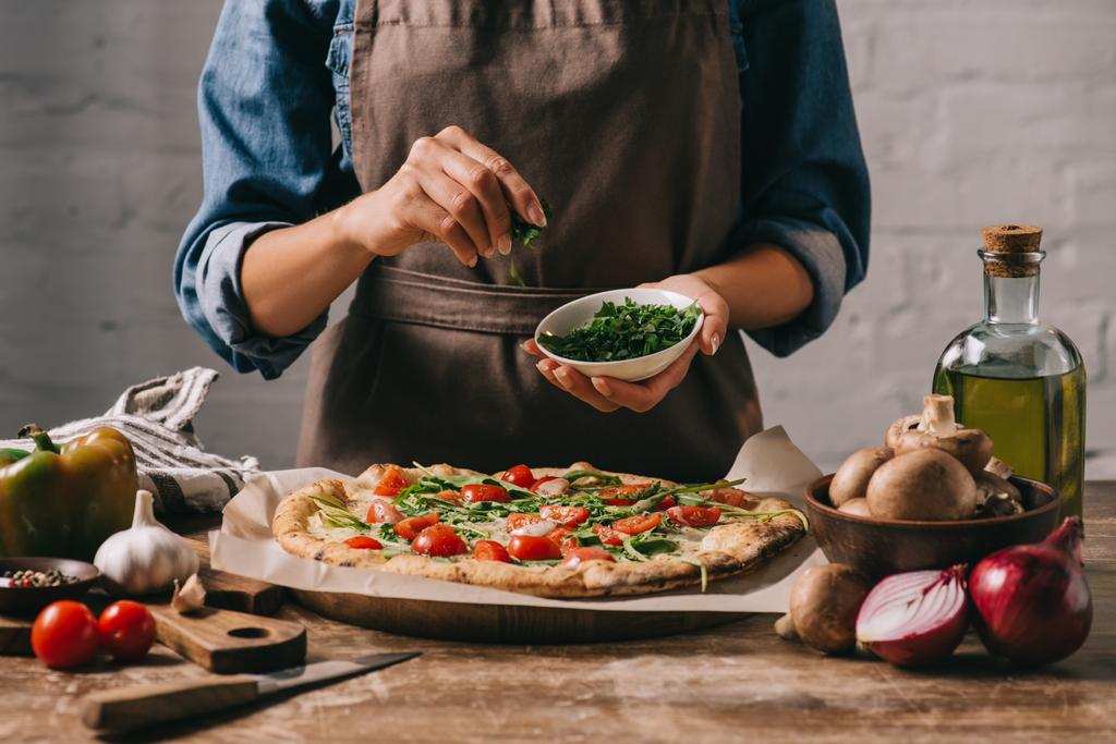 Teilansicht einer Frau in Schürze, die Petersilie auf gekochte Pizza auf Holzoberfläche gießt - Foto, Bild