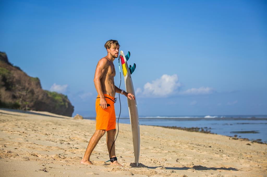 vista lateral del joven con tabla de surf en la playa de arena cerca del océano
 - Foto, imagen