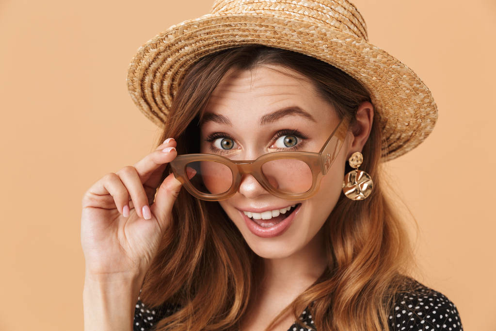 Portret van vrolijke jonge vrouw 20s dragen strooien hoed en zonnebril camera geïsoleerd op beige achtergrond kijken - Foto, afbeelding