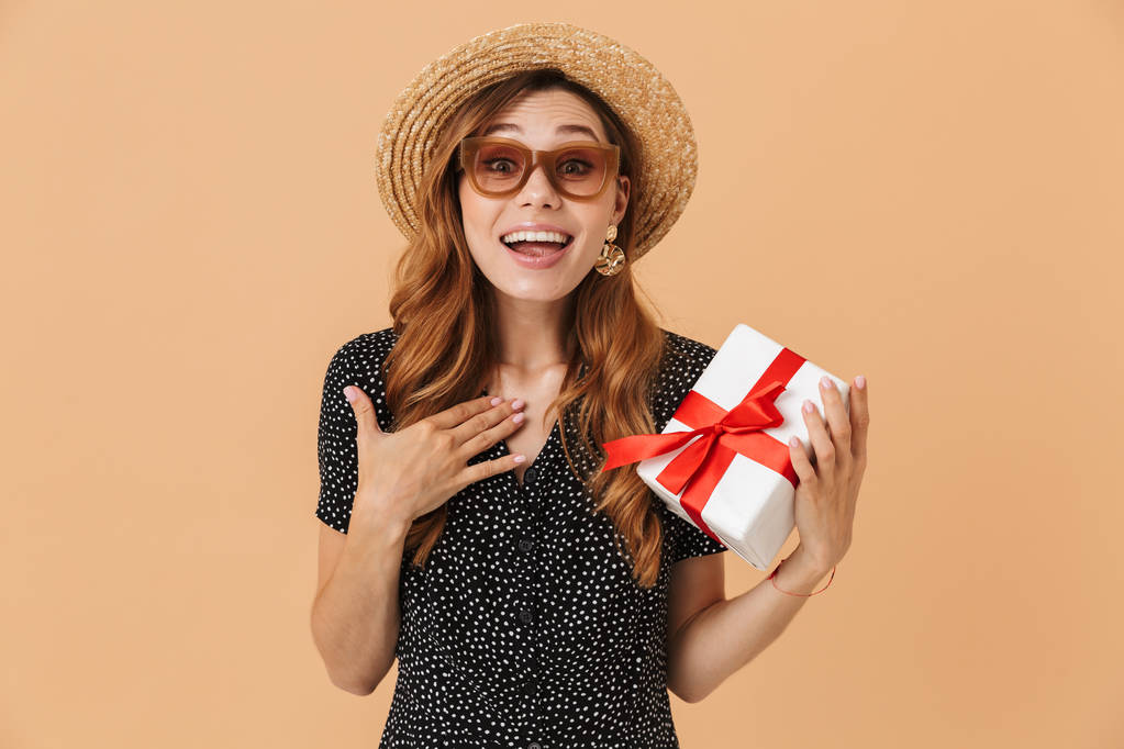 Retrato de mulher bonita surpreso 20s vestindo chapéu de palha e óculos de sol sorrindo enquanto segurando caixa de presente com fita vermelha isolada sobre fundo bege
 - Foto, Imagem