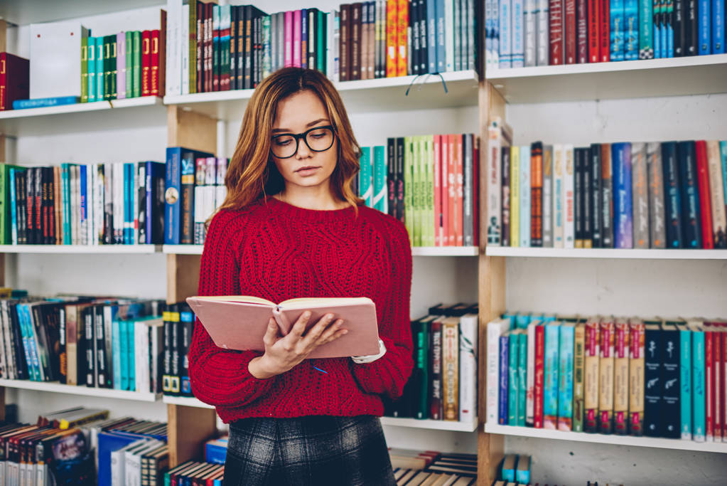 Sprytna kobieta dziennikarz wyszukiwanie informacji dla artykułu stojąc z literatury z półki na tle, atrakcyjna młoda kobieta w okulary czytanie bestsellerem w college university - Zdjęcie, obraz