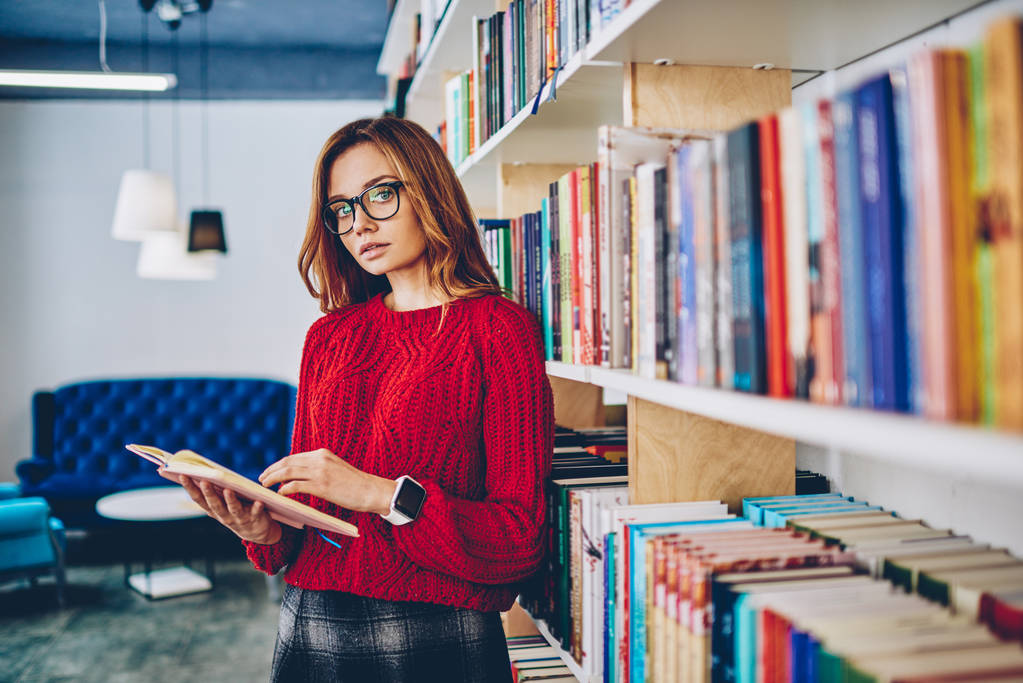 Portret połowa długości uroczy hipster dziewczyna w okulary do korekcji wzroku Podaj spędzać czas na literatura hobby stojącego obok półki na książki w bibliotece, koncepcji, wiedzy i edukacji - Zdjęcie, obraz