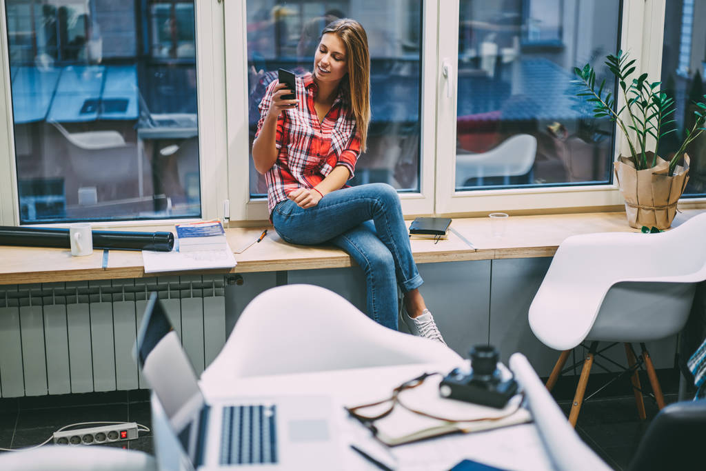 Positif caucasien jeune femme habillée en tenue décontractée assis sur le rebord de la fenêtre et la lecture de la notification entrante sur l'appareil smartphone en utilisant une connexion Internet 4G dans le studio moderne pendant la pause
 - Photo, image