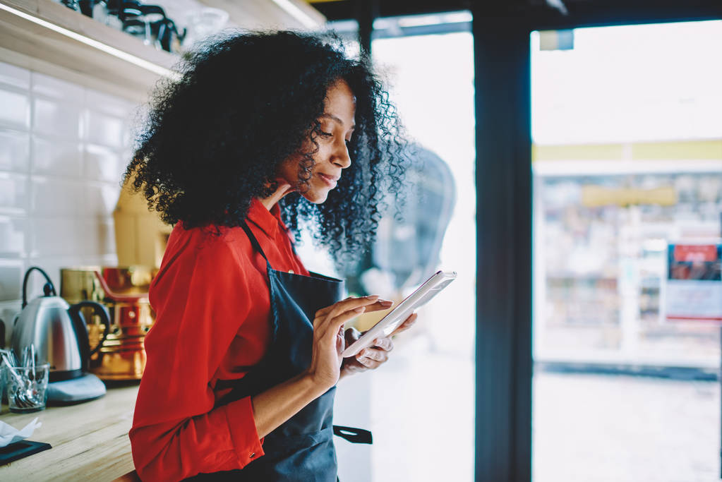 Μερική άποψη του επαγγελματική αφρικανική αμερικανική σερβιτόρα σε ενιαίο έλεγχο της παραγγελίας του πελάτη σε στέκεται συσκευή pad μοντέρνα πινελιά στο χώρο εργασίας σε καφέ. Συλλογισμένος σκούρο δέρμα barista πληκτρολογώντας κείμενο στο tablet - Φωτογραφία, εικόνα