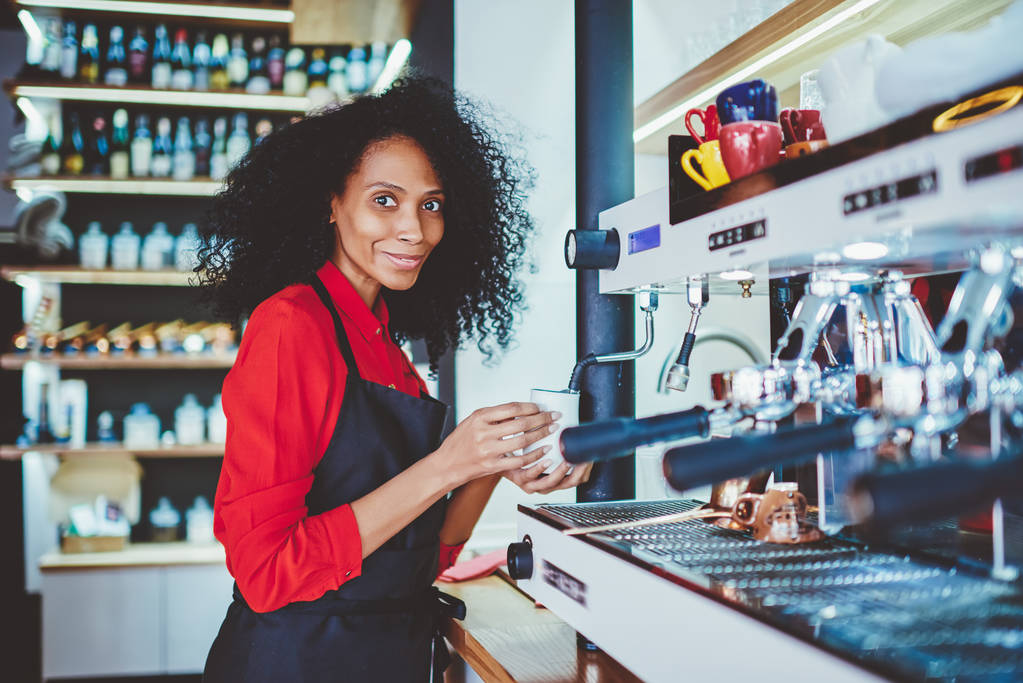 Πορτρέτο του θετική Αφρικανικός Αμερικανός βιώσει barista στην μαύρη ποδιά χαμογελώντας σε φωτογραφική μηχανή, ενώ στέκεται στην γραμμή και προετοιμασία καφέ το ποτό σε μοντέρνα μηχανή στο δικό café ανάπτυξη επιχειρήσεων - Φωτογραφία, εικόνα