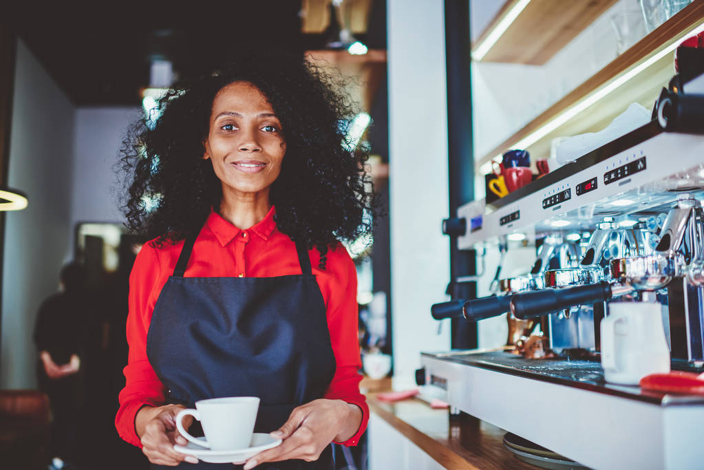 Porträt einer positiven jungen Kellnerin mit dunkler Haut, die eine Tasse Aromakaffee in der Hand hält und in der Nähe moderner Maschinen steht. - Foto, Bild