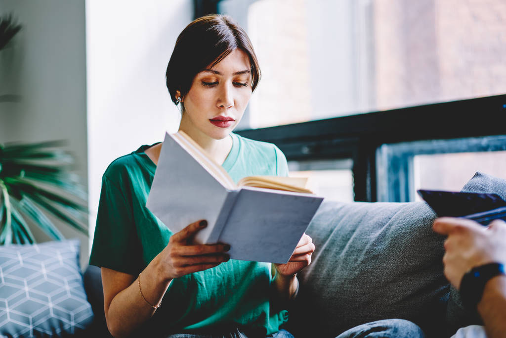 konzentrierte junge Frau liest Literaturhandlung in Buch, bereitet sich auf bevorstehende Prüfung vor und studiert zu Hause mit modernem Interieur. Nachdenkliches Hipstermädchen genießt Bestseller in der Wohnung in der Freizeit - Foto, Bild