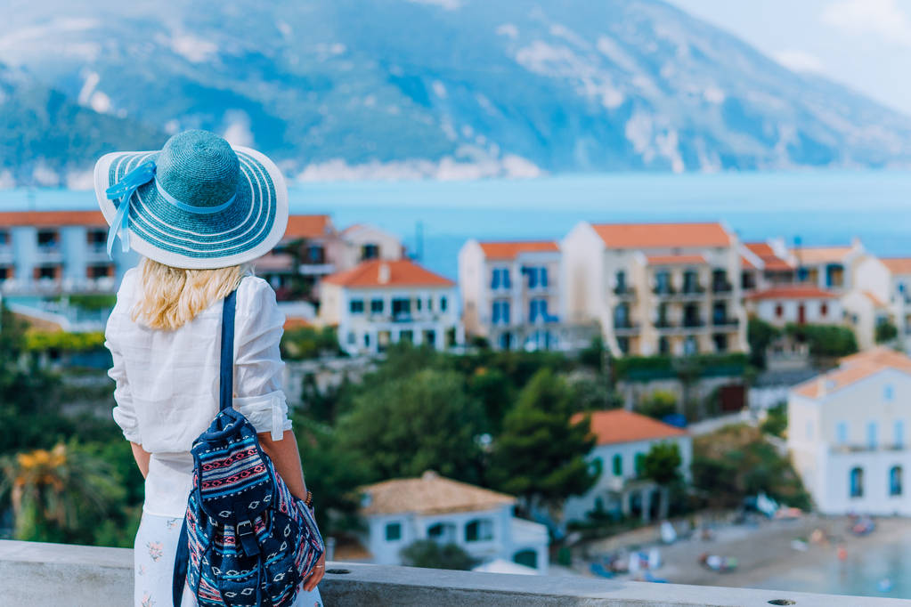 Mladé stylové turistické žena těší pohled na barevné klidné vesnice Assos. Modelka na sobě Modrý klobouk, bílé oblečení a cestovní batoh těší její letní dovolená v Kefalonia, Řecko - Fotografie, Obrázek