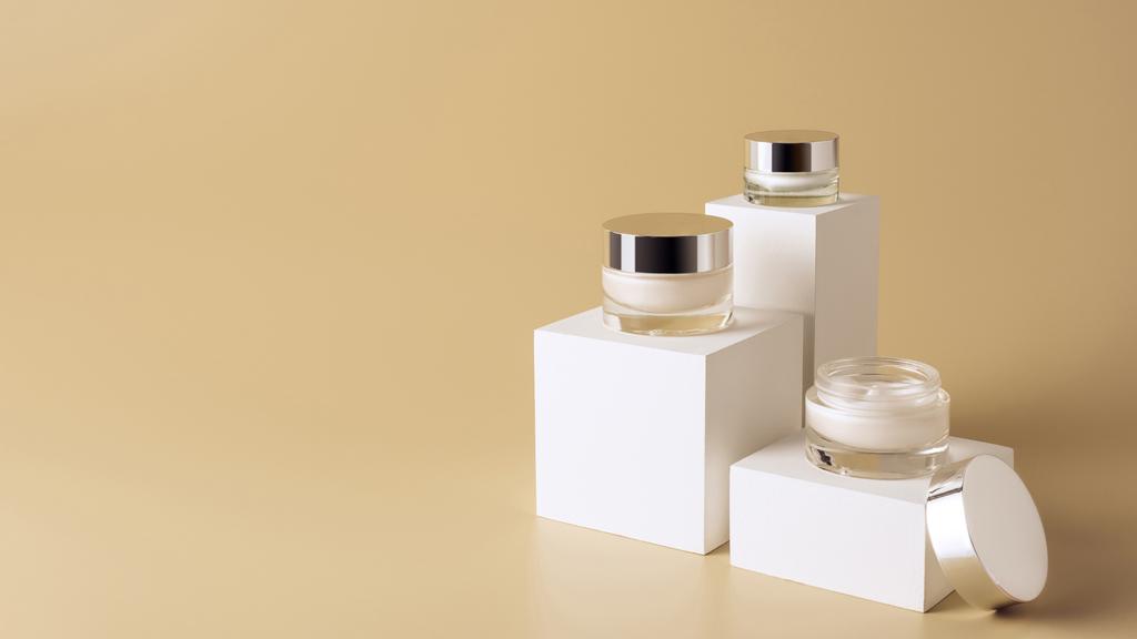 Vista de cerca de las cremas faciales y corporales en frascos de vidrio sobre cubos blancos sobre fondo beige
 - Foto, Imagen