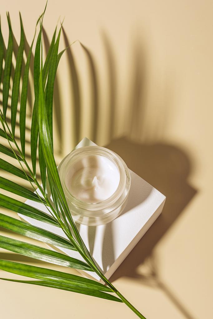 close up view of moisturizing cream in glass jar with palm leaf on beige background - Photo, Image