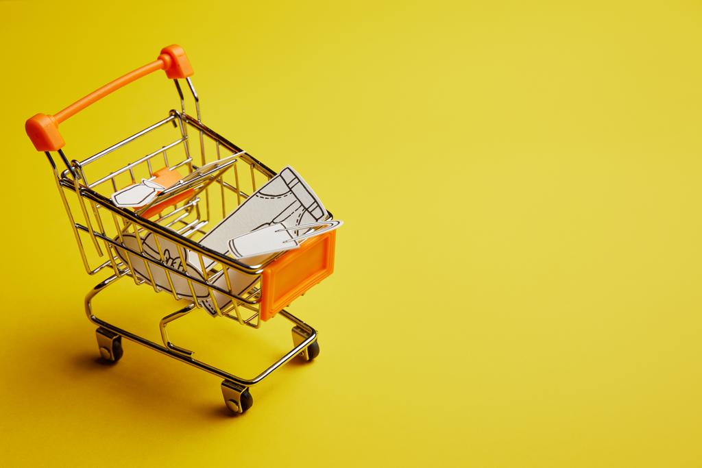 Nahaufnahme eines kleinen Einkaufswagens mit Kleidung aus Papier auf gelbem Hintergrund - Foto, Bild