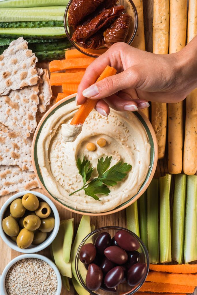 partial view of woman and arranged hummus in bowl, pita bread, cut vegetables, dried tomatoes and olives - Photo, Image