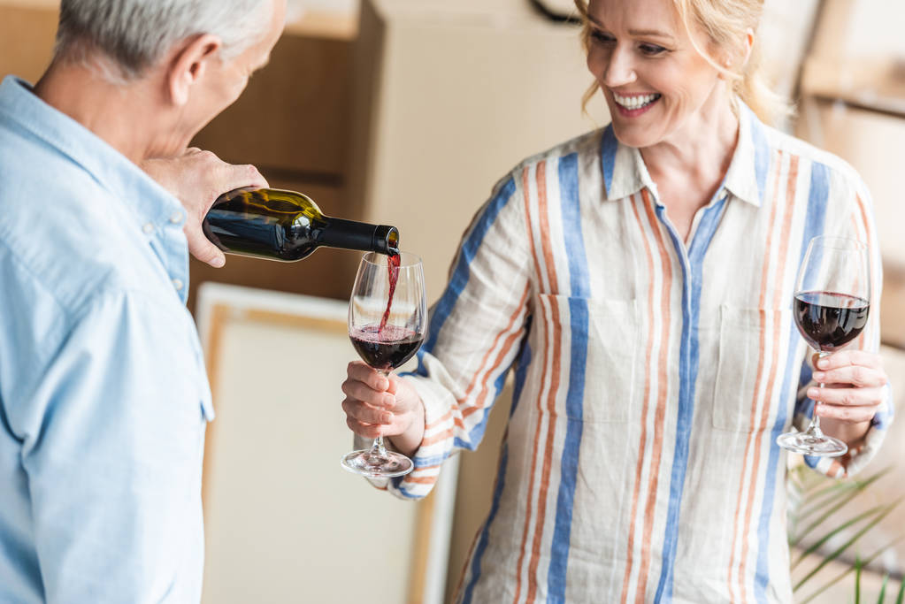 Schnappschuss eines Mannes mit grauen Haaren, der seiner glücklichen Frau beim Umzug in ein neues Haus Wein einschenkt  - Foto, Bild