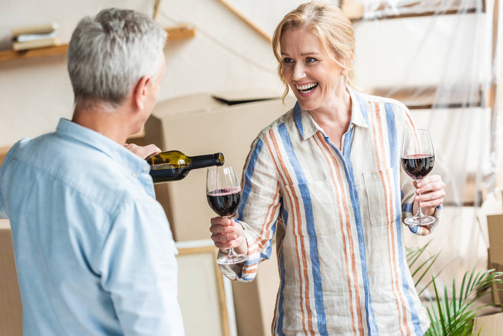 Senior man gieten van wijn tot gelukkige vrouw terwijl verhuizen in nieuw huis   - Foto, afbeelding