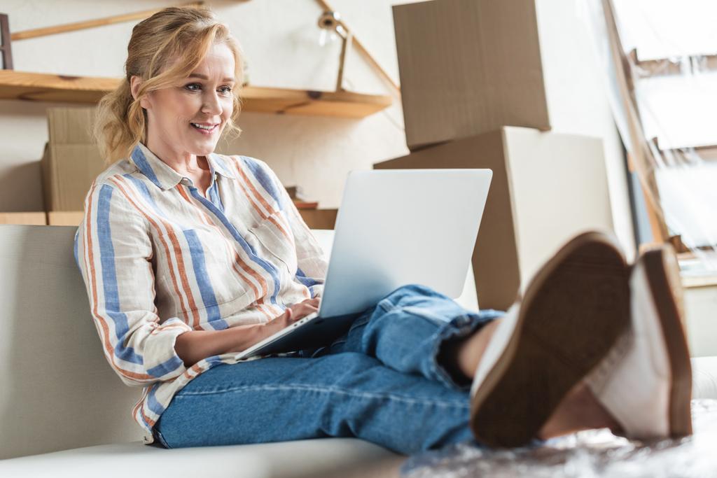 красива усміхнена зріла жінка використовує ноутбук і сидить на дивані в новому будинку
 - Фото, зображення