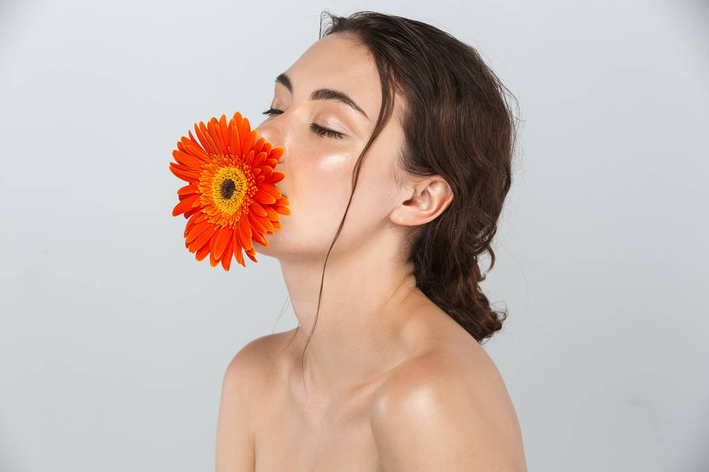 Szépség make-up jelentenek az arcán piros gerbera virág vonzó fiatal topless nő portréja elszigetelt, felett szürke háttér - Fotó, kép