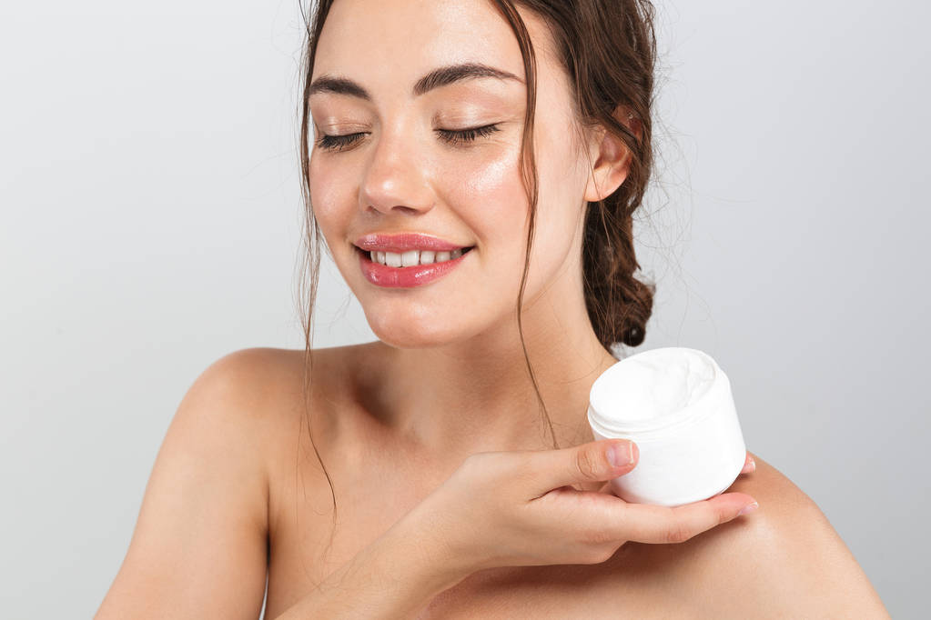 Retrato de belleza de una mujer en topless bastante joven con contenedor de sujeción de maquillaje con crema corporal aislada sobre fondo gris
 - Foto, imagen